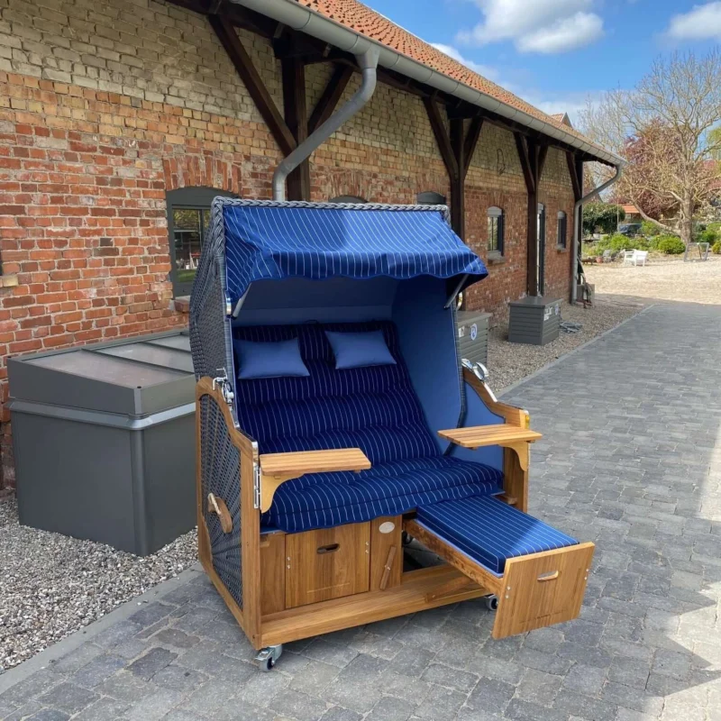 Strandkorb Prieß, Modell Meister mit Iroko Holz und dunklem PE-Geflecht, blau gestreift, in einem Garten setting.