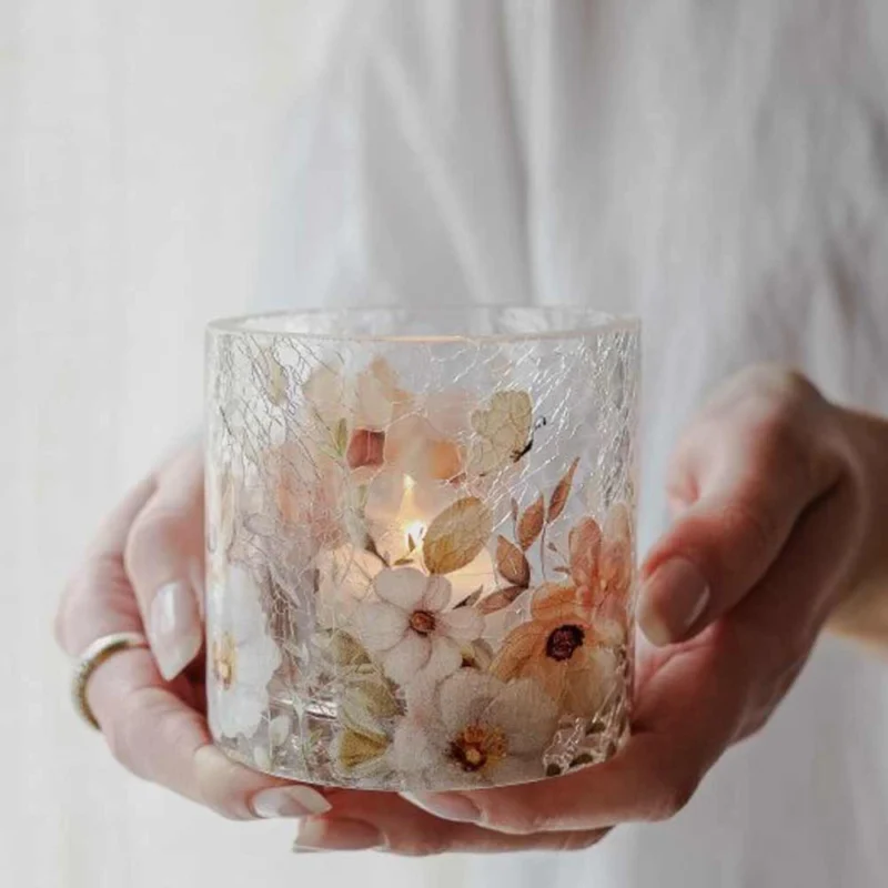 Eine Person hält den Glaskerzenhalter „Majas Cottage – Windlicht Blossom & Butterflies“, der mit gepressten Blumen und Schmetterlingen geschmückt ist und in dem sich eine brennende Kerze befindet.